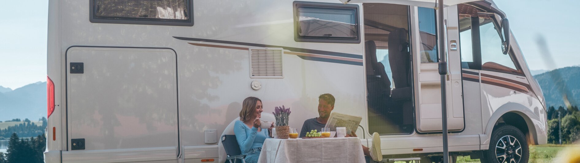 Ein Mann und eine Frau sitzen vor ihrem Campervan unter dem Sonnenschirm und trinken Kaffee.